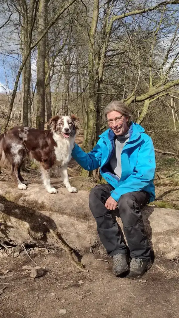 Dett Heidkamp sitzt lächelnd mit einem Hund auf einem Baumstamm im Wald